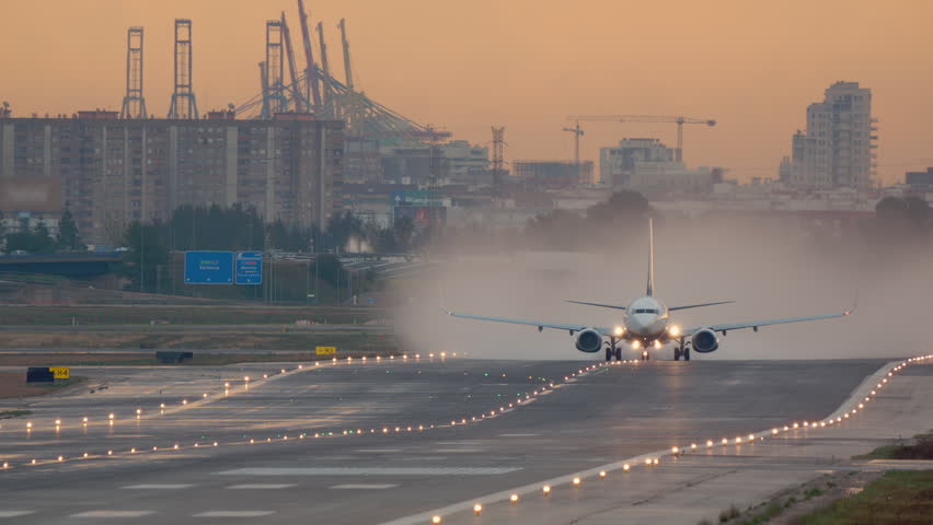 Twin engine passenger jet throwing a cloud of mist and vapor behind it, while moving along the runway during a warm orange sunrise at a coastal airport