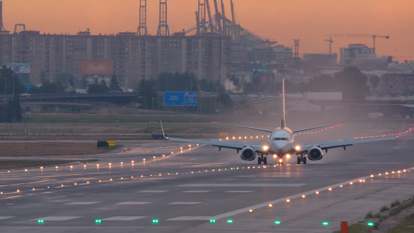 Narrow body passenger jet rolling head on along the runway under a glowing orange sunrise sky with port cranes and city buildings on the horizon