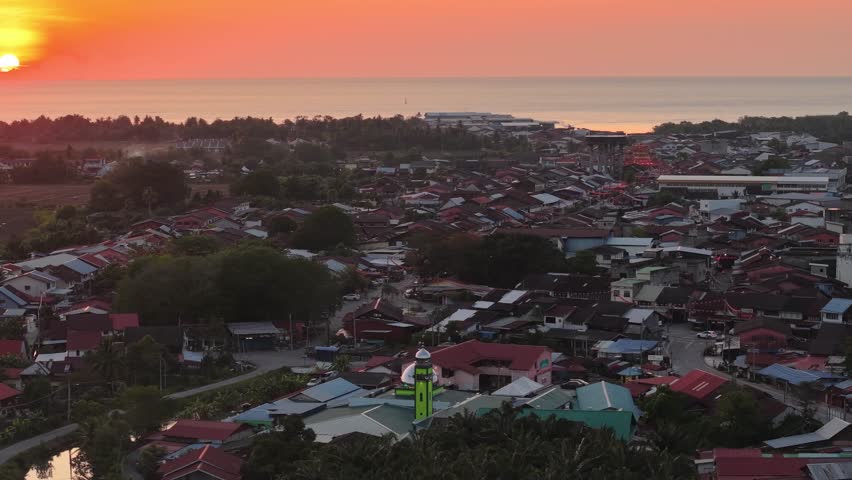 Warm orange sunset over Tanjung Piandang coastline and village from drone, mosque towers among rooftops.