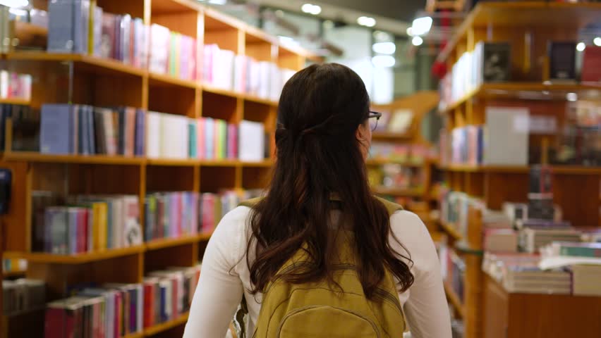 Latin Female Student with Glasses and Backpack Walking in Bookstore Library Aisle Back View Medium Shot
