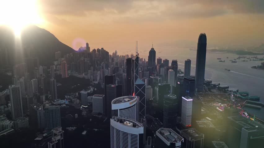 Harbor evening vista at golden hour illuminating skyline and ships at dusk in Hong Kong. Sunset peak flare over harbour filmed by drone. Light spilling across towers and coastline ridge silhouettes