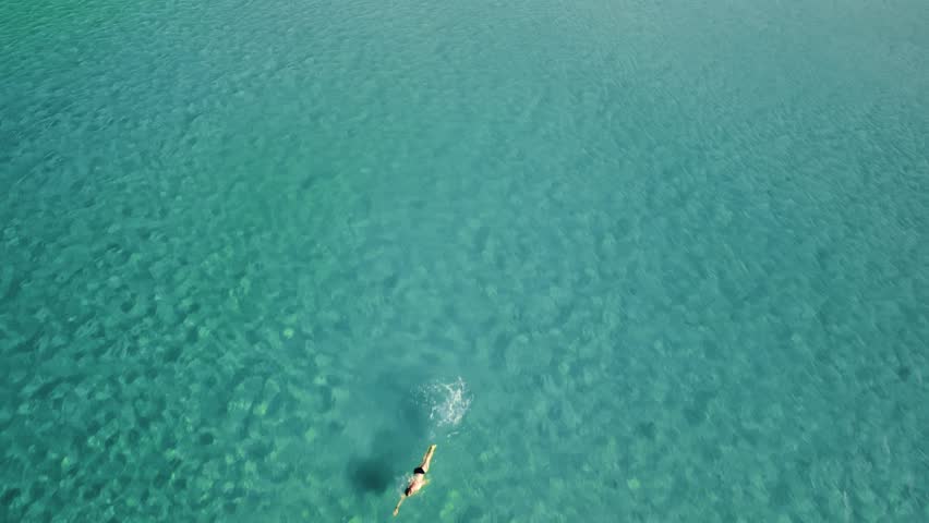 Drone captures man swimming in bright shallow water environment. Aerial view showcasing male holiday maker exercising and coordinating in sunlit shallow coastal waters. Travelling and relaxing theme