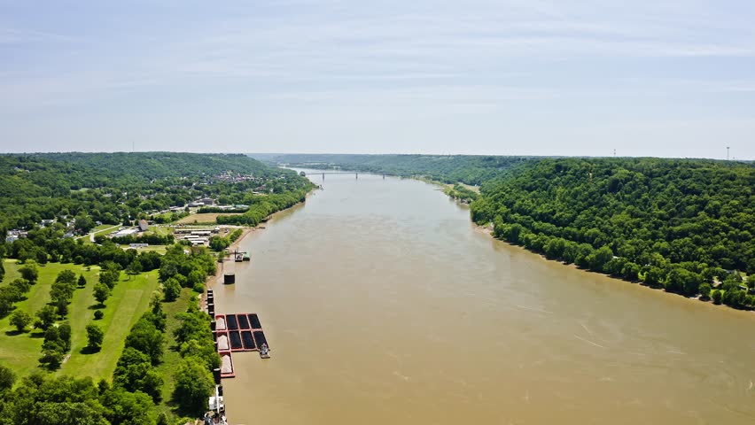 Drone view of the Ohio River near Madison, Indiana, with forested hills, riverfront landscape, barges, and small communities along the shoreline on a clear summer day.