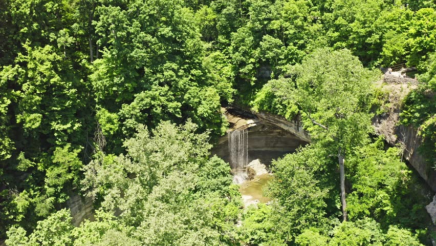 Drone view of Tunnel Falls surrounded by lush summer forest in Clifty Falls State Park, Indiana. The waterfall cascades over layered rock formations into a scenic gorge within one of southern Indiana