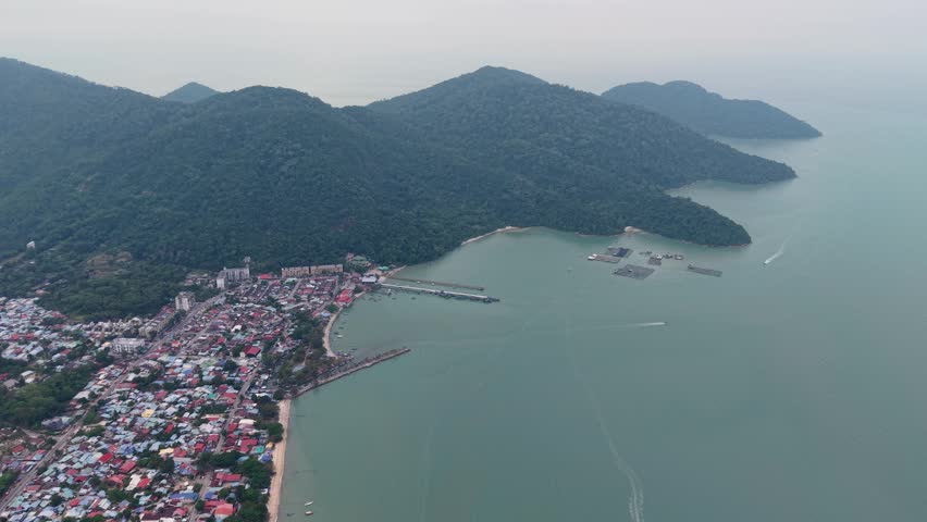 Drone footage showing Teluk Bahang town, pier, coastline, forested hills.