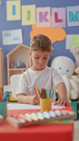 Boy writing with a pencil at a red classroom table with colored pencils and xylophone inside a school building; quiet concentration.