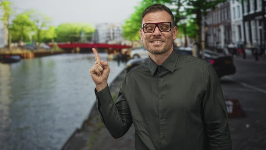 Man wearing glasses points finger toward canal and red bridge while standing on a cobblestone street near parked cars; playful confidence.