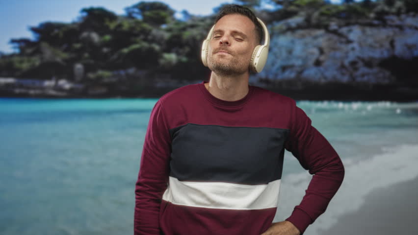 Man wearing headphones, hand on hip and head tilted while listening at a beach in a maroon sweater; confidence leisure.