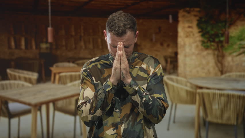 Man, soldier in camouflage uniform, with hands clasped in prayer, looking upward in a rustic dining building; devotion reflection.