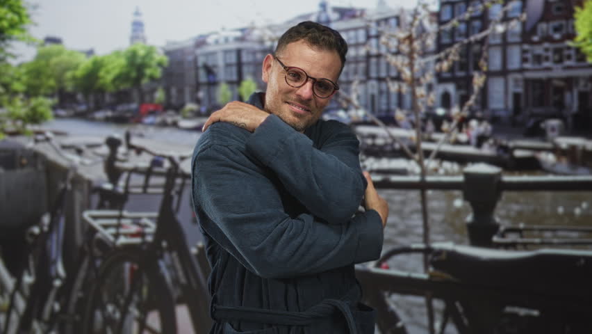 Man in robe hugs his arms across chest beside parked bicycles on a canal backdrop in a studio; melancholy.
