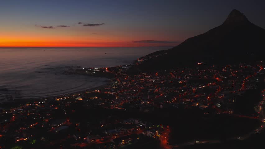 Aerial twilight view of Cape Town, South Africa, shows a crescent bay, rocky shore, amber city lights, gentle waves, vessels, and a dark hillside at sunset.