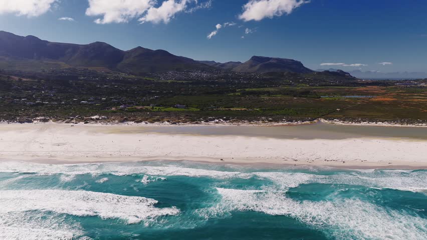 Aerial view of a long white sand beach near Cape Town, South Africa, with surf, dunes, scrub, houses, and mountains. Tiny figures and vehicles appear in bright daylight.