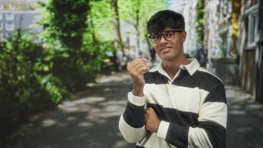 Man touching chin with hand, wearing glasses and black and white striped rugby shirt, gesturing with open palm on leafy street; thoughtful contemplation.