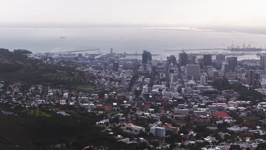 Aerial view of Cape Town, South Africa, shows downtown towers, Table Bay, breakwaters, container cranes, docked ships, and V and A Waterfront in soft late day haze.
