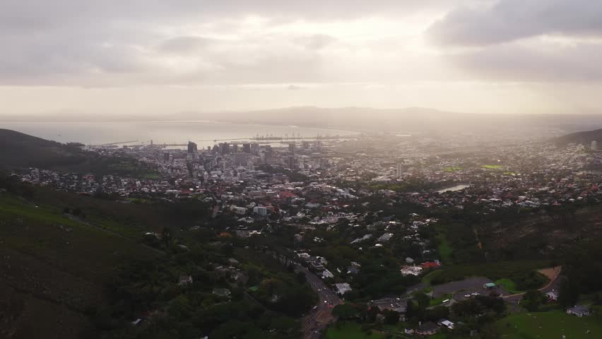 Aerial panorama of Cape Town shows downtown skyline, Table Bay, harbor cranes, and Table Mountain slopes. Stormy sunrays backlight city, roads and traffic wind downhill.