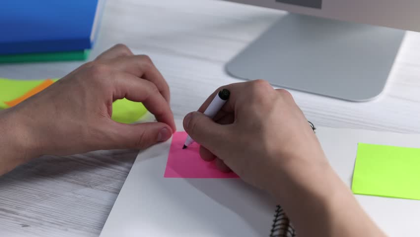 Man writing word Task on pink sticky note at light wooden table, closeup. Planning and deadline