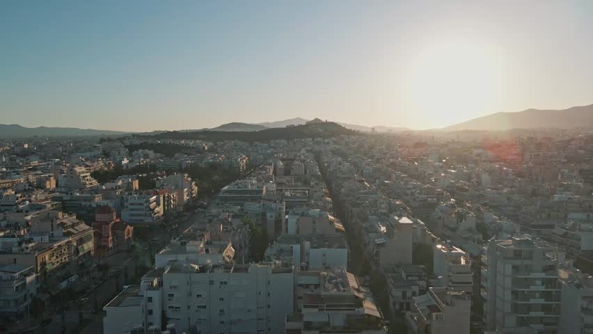 Aerial view of the dense urban landscape with rooftops and city streets under a golden sunset sky in Athens, Greece.