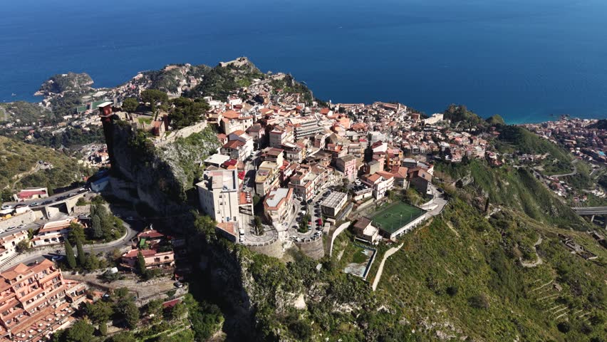 Aerial view of the hilltop village of Castelmola with historic stone buildings and winding roads overlooking the blue sea in Castelmola, Sicily, Italy.
