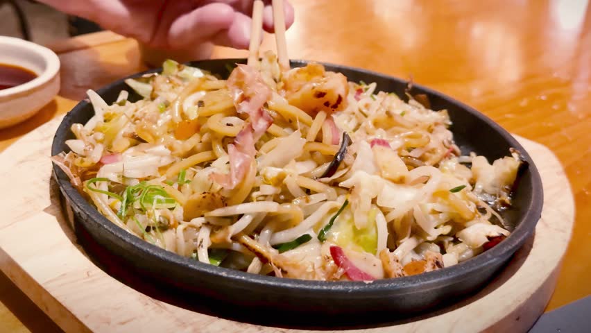 Chopsticks lift steaming Japanese yakisoba noodles with shrimp and bonito flakes from a skillet.
