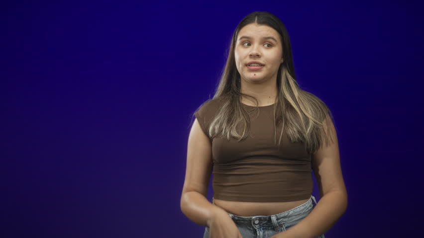 Young woman palms up shrug showing exposed midriff, smiling and gesturing with open hands while wearing a brown crop top and jeans in studio; casual uncertainty.