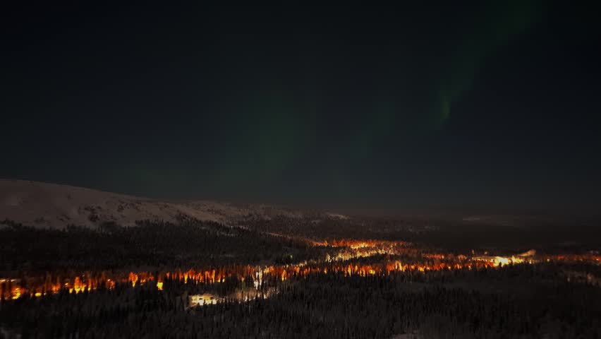 Aerial view of the aurora borealis dancing over the snow-covered forest and illuminated village of Luosto in Sodankyla, Lapland, Finland.