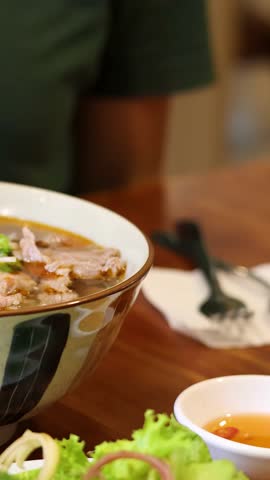 A close-up shot shows a person lifting rice noodles from a bowl of beef pho.