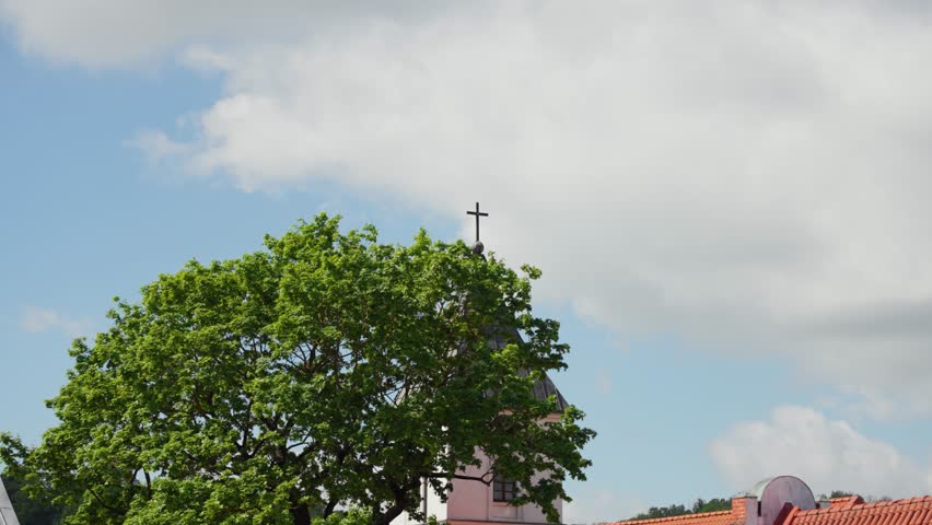 Wide shot of a church steeple topped with a cross, partly obscured by a leafy green tree and red tiled rooftops under a bright blue sky with fluffy white clouds. Vibrant, natural daylight.