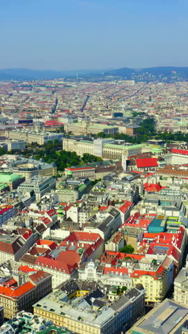 Vertical video. Vienna, Austria. The historic center of the city spreads below in a sweeping aerial panorama, revealing a dense tapestry of terracotta rooftops, Baroque church spires, and grand imperial palaces that define one of Europe