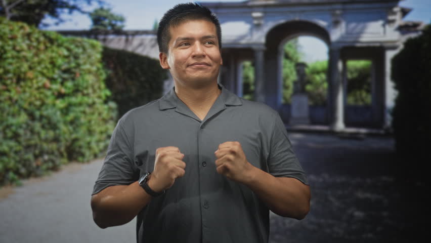 Man in gray shirt with clenched fists and visible forearms standing before an arched building entrance, looking up; calm confidence.