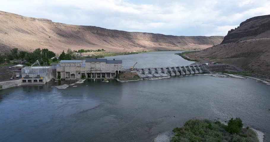 Aerial view of a dam in a desert landscape. Calm water fills the reservoir. Mountain ranges frame the scene.
