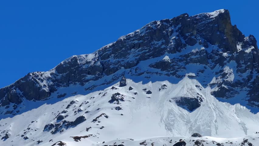 Aerial view of a rugged snow-capped mountain peak with rocky cliffs under a clear blue sky in Grisons, Switzerland.