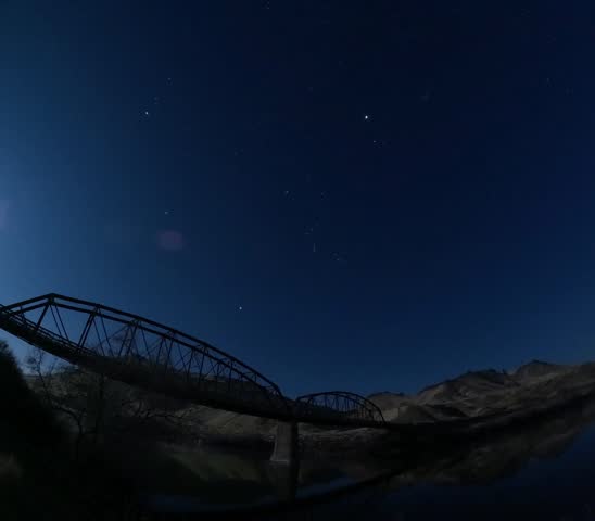 Dark night sky glows with a bright moon. Star trails swirl across the blue sky. Metal bridge spans a still river.