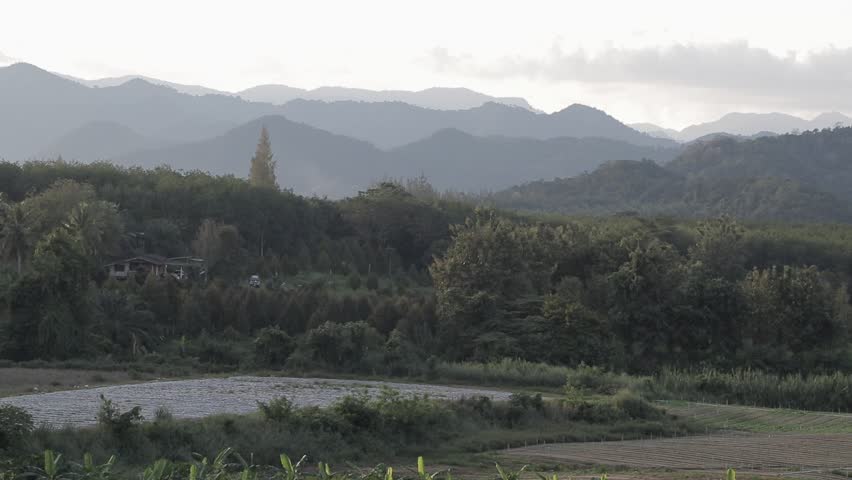 A scenic panoramic view of rural agricultural fields and farming land nestled against a lush tropical forest and majestic mountain range.