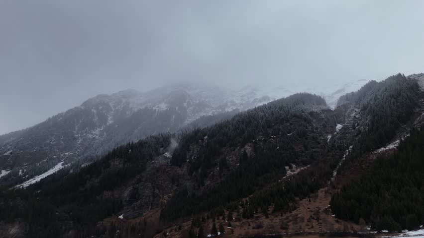 Cinematic lateral drone flight gliding above a snow-covered pine forest in South Tyrol, Italy, with fog weaving through the treetops. The camera moves smoothly sideways, revealing the dense forest below and snow-capped Alpine mountains emerging in the distance under a gray overcast sky. Mist and low clouds add depth and a moody, atmospheric quality, emphasizing the scale of both forest and mountains. Perfect for cinematic aerials, winter alpine landscapes, foggy forest explorations, and immersive nature storytelling.
