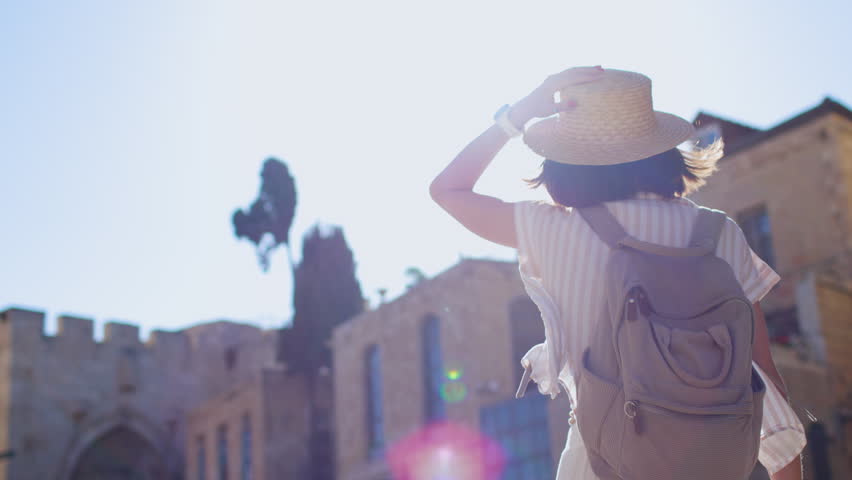 Rear view of a young woman with dark shoulder-length hair and a backpack crossing a sunlit Jerusalem city square, with sunlit buildings and a soft blur in the background. Jerusalem. Israel