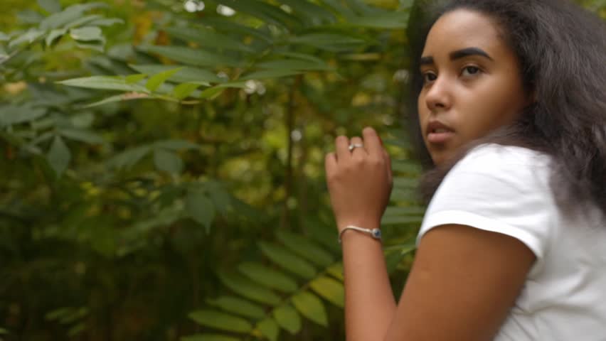 Pensive young woman with curly hair walking and posing outdoors on a stone path surrounded by trees.