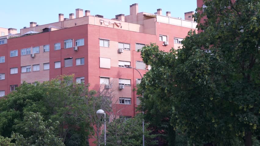 A daytime medium shot of a multi-story red brick residential apartment building in Madrid, Spain. The building features numerous windows, shutter blinds, and air conditioning units on the facade, partially framed by lush green trees and bushes in the foreground under a clear sky.