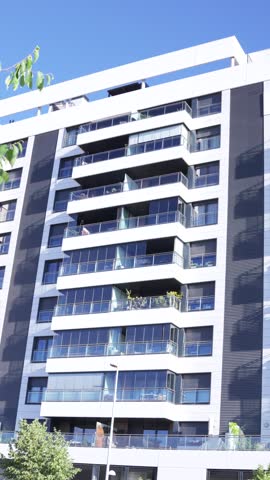 Vertical view of a modern residential apartment building with glass balconies and contemporary urban architecture under a clear blue sky in Madrid, Spain.