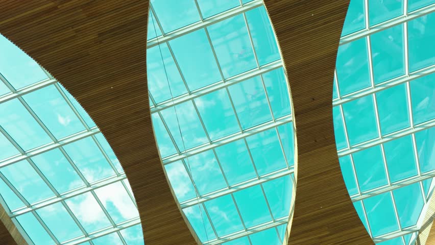 Curved skylight ceiling inside retail center with moving clouds visible through glass panels. Futuristic architectural pattern combining transparent roof design daylight and spacious shopping