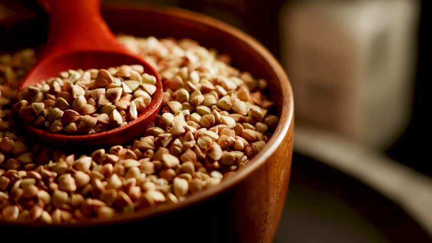 Organic green buckwheat grains release thick white vapor from a dark bowl. An orange scoop rests amongst the healthy food, ready for serving.