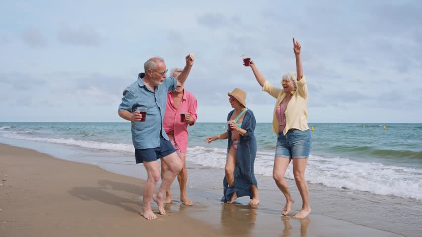 Cheerful group of elderly friends happily dancing and laughing together on the seashore. Four retired people celebrating and having fun on vacation