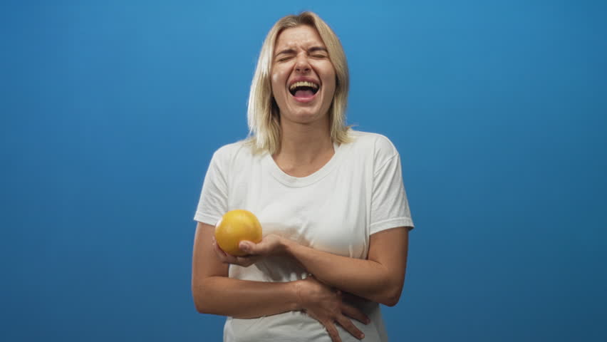Young blonde woman holds an orange in one hand while laughing and clutching her stomach in a blue studio; amusement.