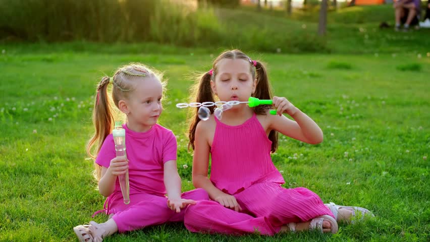 children blowing bubbles in the park in summer, two girls walking and having fun on a green lawn, summer holidays