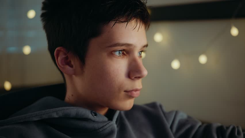 A focused teenage boy with blue eyes looking at a screen in a dark room. Bokeh lights are visible in the background.