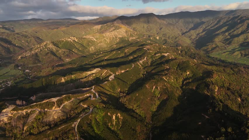 Rugged mountain range with winding dirt tracks carved into golden hillsides, layered ridges and forest valleys bathed in warm sunrise light under dramatic clouds. New Zealand. Aerial backwards wide