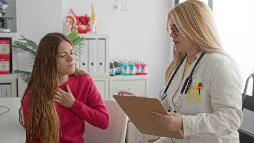 Two women doctor and patient, patient hand on chest while doctor with stethoscope reads clipboard in building; concern medical consultation.