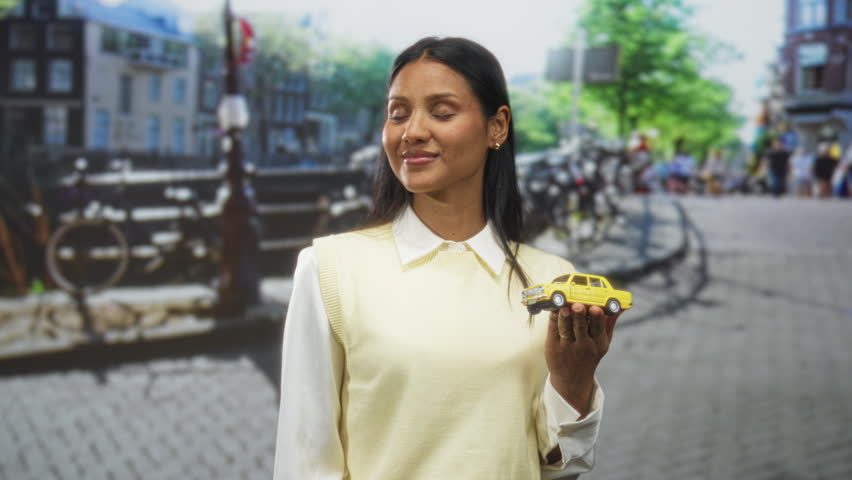 Woman holding yellow toy car in right hand on cobbled street with bicycles and canal behind her; nostalgia travel.