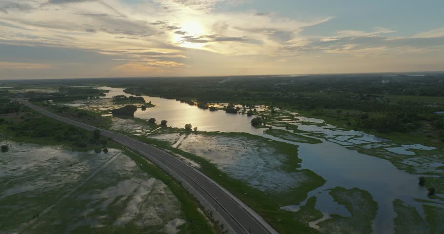 Cinematic aerial drone footage of a winding road passing through a tranquil landscape with a small lake between the roadway during golden hour. Vibrant sunset colors illuminate the sky.
