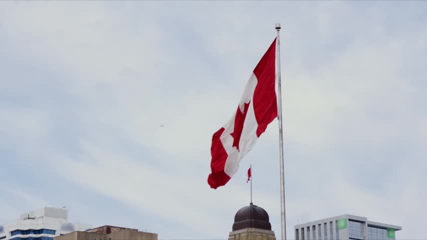 The Canadian flag is flying in downtown Halifax, Nova Scotia. People walk nearby as the flag moves with the wind against the cloudy sky