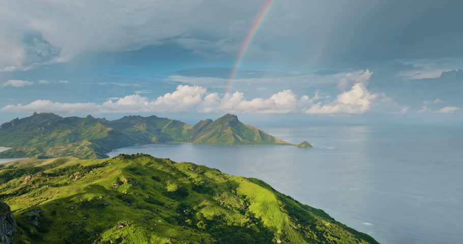 Green mountains and lush tropical landscape of Fiji Islands with a colorful rainbow stretching across the sky, overlooking bays and vast Pacific Ocean under a cloudy sky. Wild nature aerial landscape